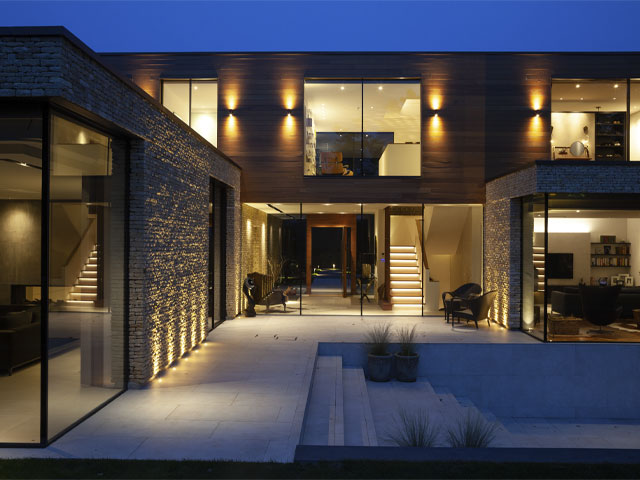 a modern self-build with large windows and cladding of stone and timber lit up at night 