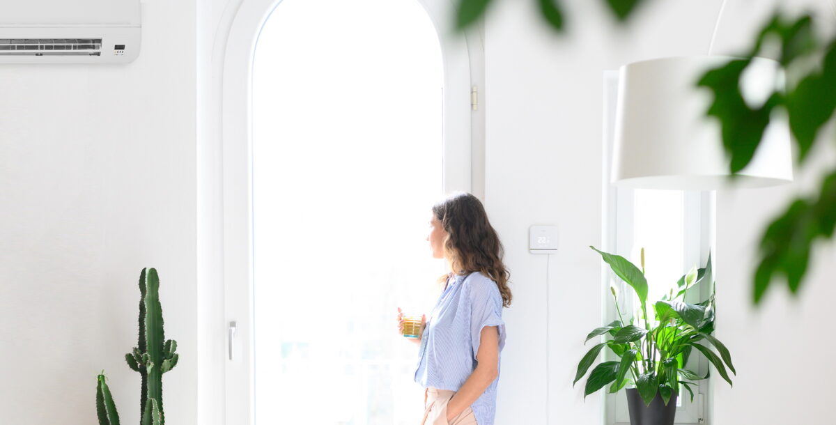 Lifestyle photo of a woman looking out of the window, between the Tado Smart Air Conditioning Controller and a wall-mounted air conditioner